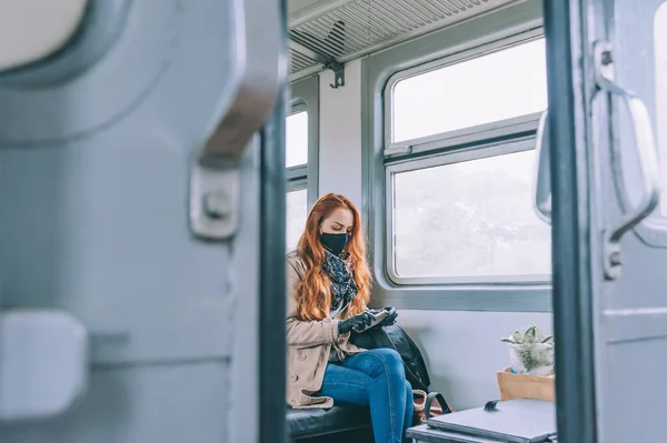 赤い髪の少女は 彼女の手にスマートフォンを保持し 黒い医療マスクと 電車の馬車に座っている — ストック写真