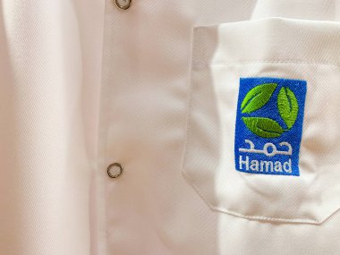 Arapça ve İngilizce Hamad logolu bir doktor veya asistan beyaz iş elbisesi. Hamad (Hmc) en büyük sağlık kuruluşudur. Nisan 2019, Doha/ Katar.