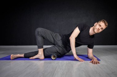 Genç adam yoga silindirinin yardımıyla dış kalça kaslarını esnetiyor..