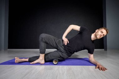 Genç adam yoga silindirinin yardımıyla dış kalça kaslarını esnetiyor..