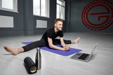 Genç, atletik adam dizüstü bilgisayarın önünde geriniyor. Online yoga dersi.