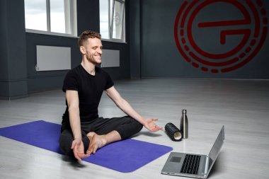 Genç, atletik adam dizüstü bilgisayarın önünde meditasyon yapıyor. Online yoga dersi.