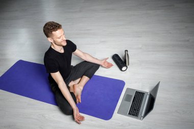 Genç, atletik adam dizüstü bilgisayarın önünde meditasyon yapıyor. Online yoga dersi.