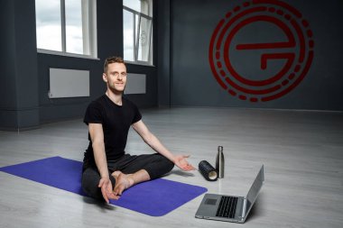 Genç, atletik adam dizüstü bilgisayarın önünde geriniyor. Online yoga dersi.