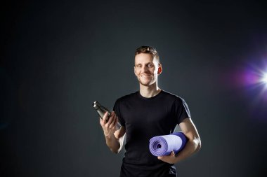 Siyah arka plandaki genç adam fitness şişesi ve spor paspası tutuyor..