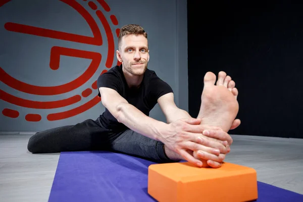 Young man is stretching his leg on yoga mat with yoga brick. Selective focus.