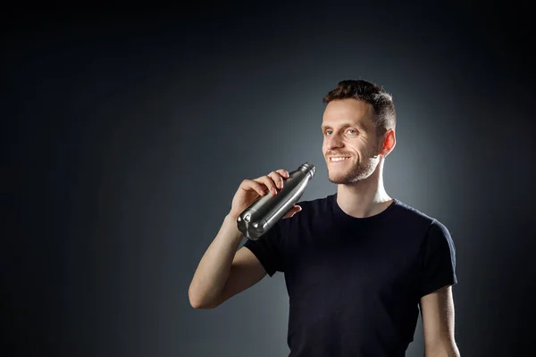 Young smiley man on the black background is drinking water from fitness bottle.