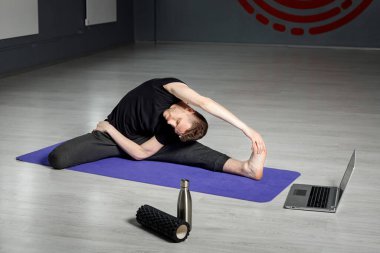 Genç, atletik adam dizüstü bilgisayarın önünde geriniyor. Online yoga dersi.