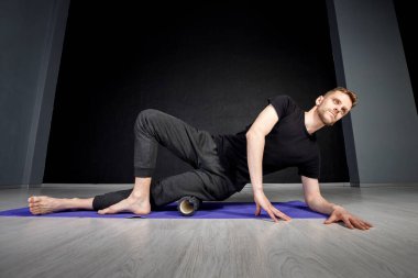 Genç adam yoga silindirinin yardımıyla dış kalça kaslarını esnetiyor.