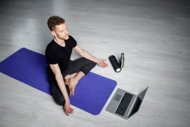 Genç, atletik adam dizüstü bilgisayarın önünde meditasyon yapıyor. Online yoga dersi.