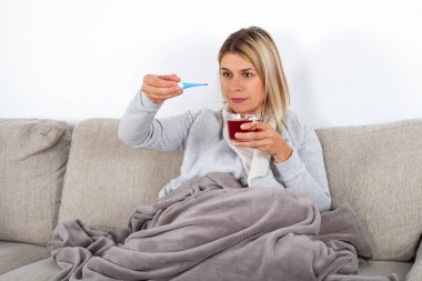 Hasta kadın sıcak bir battaniyeye sarılmış kanepede oturan bir sıcak çay ve termometre