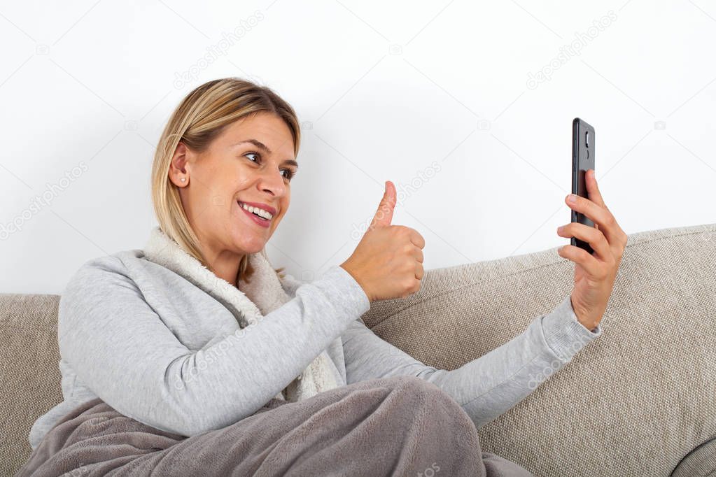 Attractive young woman talking with friend on facetime, showing thumbs up while relaxing on the sofa