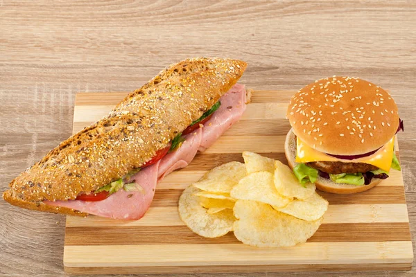 Fresh ciabatta sandwich with pork ham and veggies, delicious cheeseburger and potato chips on a wooden cutting board
