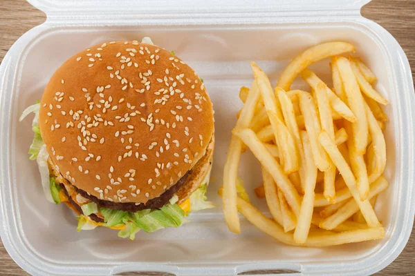 Fresh double cheeseburger and potato fries in a white plastic takeaway container on wooden background. Food delivery