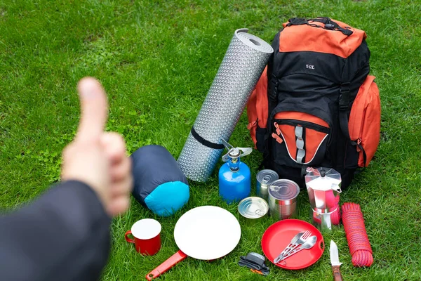 Picture of camping tools on the grass - backpack, tent, gas tank, cans, compass, etc., tourist showing thumbs up