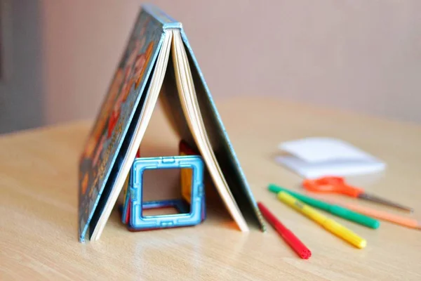 El libro se coloca en una mesa en forma de techo de una casa, forma una casa con un diseñador, junto a ella se encuentra lápices, rotuladores, tijeras, concepto de vivienda, concepto de hogar para un niño, familia . — Foto de Stock