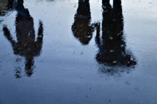 Un uomo con una mano di bambini va sotto un ombrello, riflesso sulla strada bagnata, tre figure di silhouette — Foto Stock