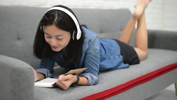 Jovem Feliz Deitada Sofá Desenhando Algo Livro Anotações Ouvindo Música — Vídeo de Stock