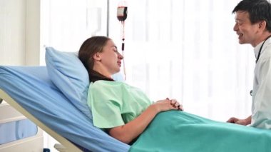 4K kıdemli Asyalı doktor beyaz hastayı yatağının başında rahatlatıyor.