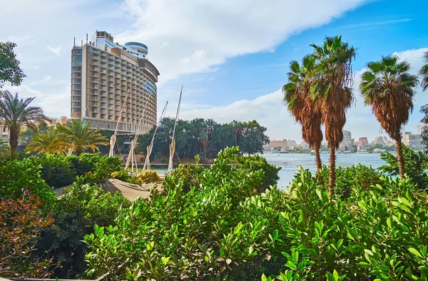 The view on modern hotel\'s building through the lush greenery of riverside garden, hiding small port with tourist feluccas, Cairo, Egypt.