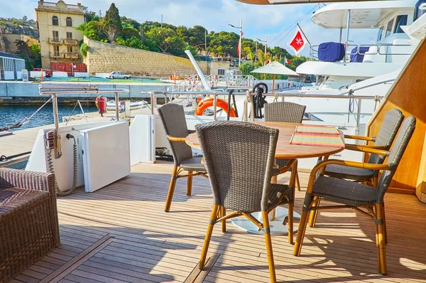 The shady terrace on the lower deck of the yacht with a table and cozy rattan chairs is the beautiful place to relax, enjoy the lunch or cup of coffee, Valletta, Malta.