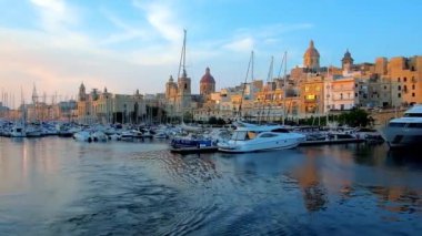 Birgu, Malta - 18 Haziran 2018: Gün batımı ışınları Ortaçağ yapılardır ve demirli yat ve tekne, Haziran 18 Birgu içinde yumuşak dalgalar üzerinde sallanan ile Vittoriosa Marina boyunca kilise aydınlatmak.
