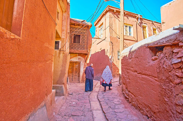 The senior couple in traditional Abyanaki clothes walks along the narrow ascent among the red adobe houses, Abyaneh, Iran.
