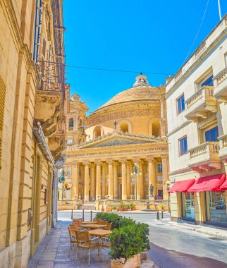 Mosta, Malta - 14 Haziran 2018: Dar gölgeli sokak açık kafe ile meşgul Rotunda meydanında görünümü ile onun ana Simgesel Yapı - Basilica varsayım of Our Lady, 14 Haziran Mosta içinde açılır.