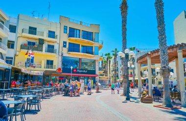 Bugibba, Malta - 14 Haziran 2018: Bay Meydanı ' nın kıyısında yer alan ana alışveriş ve gurme alan mağazaları, kafe, Bar ve palmiye altında banklar ile 14 Haziran Bugibba içinde ortasında, ağaçlar.