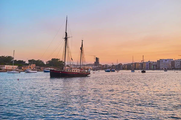 The vintage sailing ship is bobbing on the waves of Valletta Northern Harbour at the shore of Manoel Island, the evening cityscape of Sliema is seen behind it, Malta.