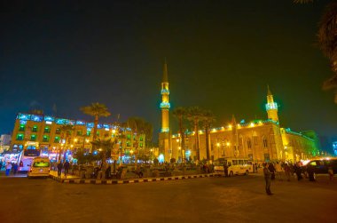 CAIRO, EGYPT - DECEMBER 20, 2017: Midan Hussein Square with illuminated Al-Hussein Mosque and neighbor edifices and spontaneous market are very crowded after dusk, on December 20 in Cairo. clipart