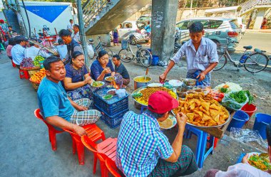 YANGON, MYANMAR - FEBRUARY 17, 2018: The crowded roadside food stalls in Chinatown with wide range of tasty local dishes with vegetables, chickpeas, deep fried cakes, on February 17 in Yangon. clipart