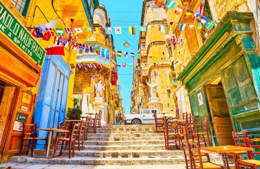 Valletta, Malta-19 Haziran 2018: St Lucia caddesinin dar inişi, 19 Haziran 'da Valletta 'da, bayrak çelençleriyle süslenmiş yerel bar ve kafelerin tablolarıyla kaplıdır.