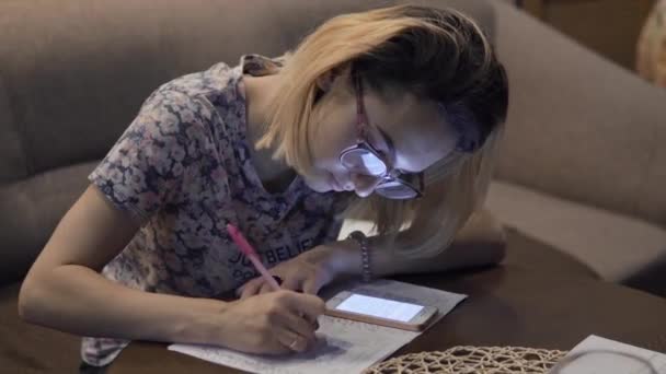 La chica escribe en las notas del cuaderno. Sentado en la sala de estar a la mesa — Vídeos de Stock