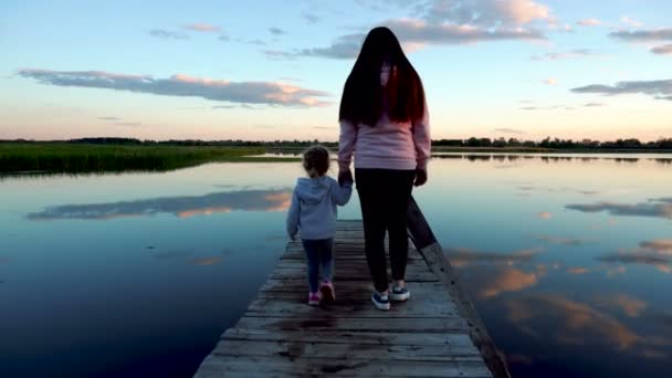 Mam en dochtertje lopen bij zonsondergang over de brug. Er is een meer rond de brug. Wolken weerspiegeld in het meer — Stockvideo