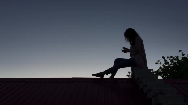 Девушка сидит на крыше с телефоном — стоковое видео