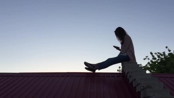 Chica sentada en el techo con el teléfono — Vídeos de Stock