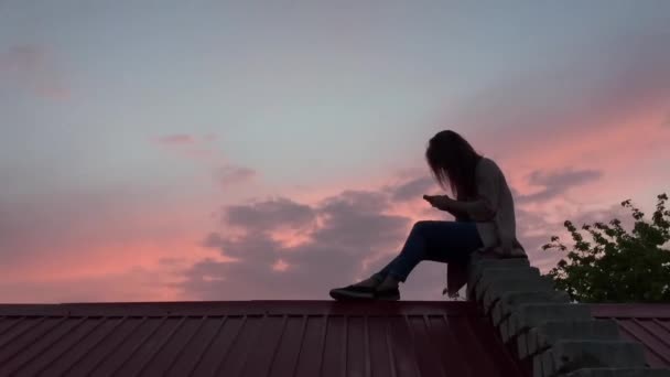 Девушка сидит на крыше с телефоном — стоковое видео