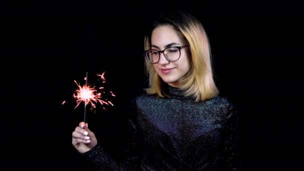 Una chica tiene una bengala en la mano. Una joven con gafas está de pie en un vestido de noche brillante sobre un fondo negro. 4k — Vídeo de stock