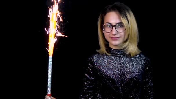 A lány tartja a tűzijáték a kezében. Egy fiatal nő szemüveg áll egy fényes estélyi ruha fekete háttér. Lassított mozgás — Stock videók