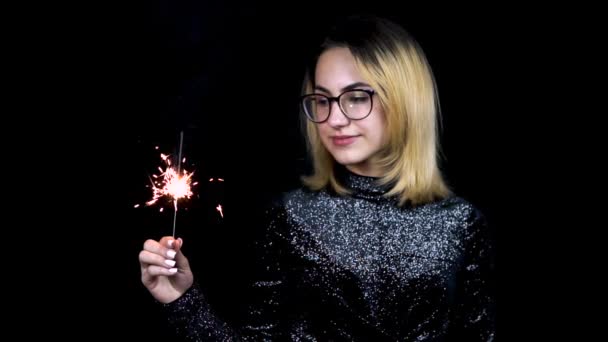 Una chica tiene una bengala en la mano. Una joven con gafas está de pie en un vestido de noche brillante sobre un fondo negro. Movimiento lento — Vídeos de Stock