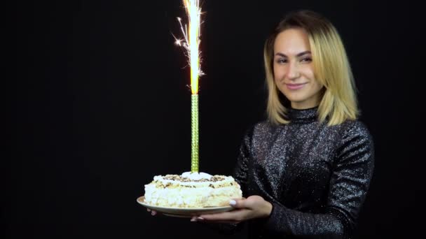 La chica sostiene un pastel de vacaciones con fuegos artificiales. Una mujer joven está de pie en un vestido de noche brillante. Fondo negro aislado — Vídeo de stock