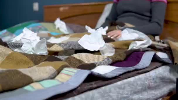 Молодые женщины чихают. Девушка лежит на кровати под пледом в кругу салфетки разбросаны. Острая респираторная вирусная инфекция — стоковое видео