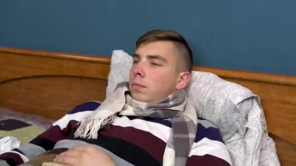 Un joven se pone un termómetro en la boca. El tipo está enfermo acostado en la cama con una bufanda alrededor de su cuello. Primer plano. — Vídeo de stock