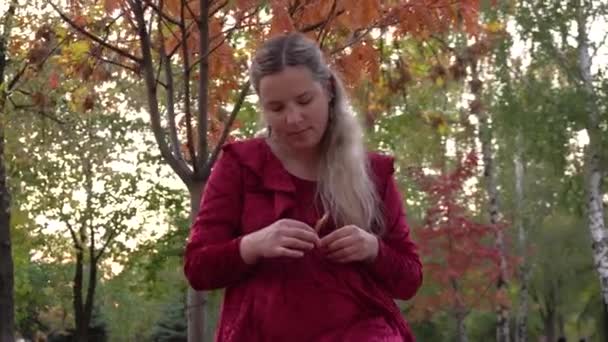 Mujer embarazada joven en un vestido rojo se sienta en el suelo bajo un árbol en un parque — Vídeo de stock