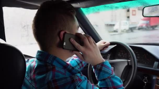 Молодой человек водит машину и говорит по телефону. Дождливая погода. Вид сзади на мужчину с заднего сиденья — стоковое видео
