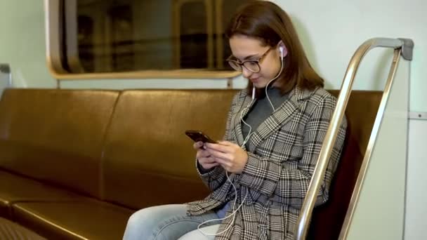 Mladá žena poslouchá hudbu na sluchátkách s telefonem v rukou ve vlaku metra. Ta dívka je v korespondenci po telefonu. Staré metro auto — Stock video