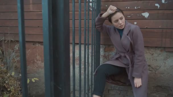 Девушка в фиолетовом пальто поставила ногу на забор — стоковое видео
