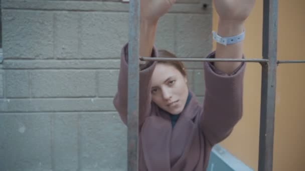 Девушка в лиловом пальто стоит за забором на улице и смотрит — стоковое видео
