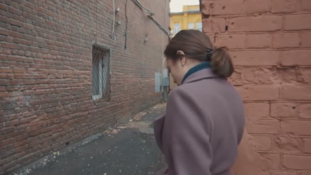 Dívka v šeříkovém kabátě s opaskem prochází uličkami mezi domy a najednou se otočí a jde dál — Stock video